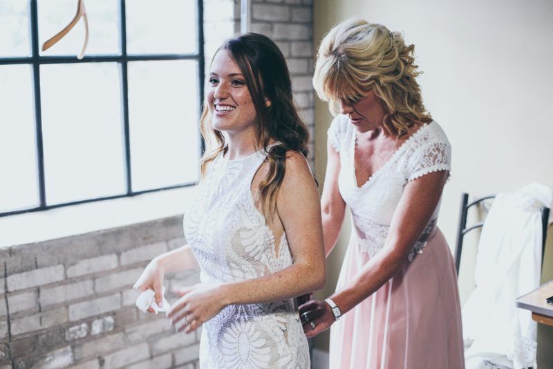 Bride smiles as a woman adjusts her wedding dress. Indoors, by a brick wall and window.