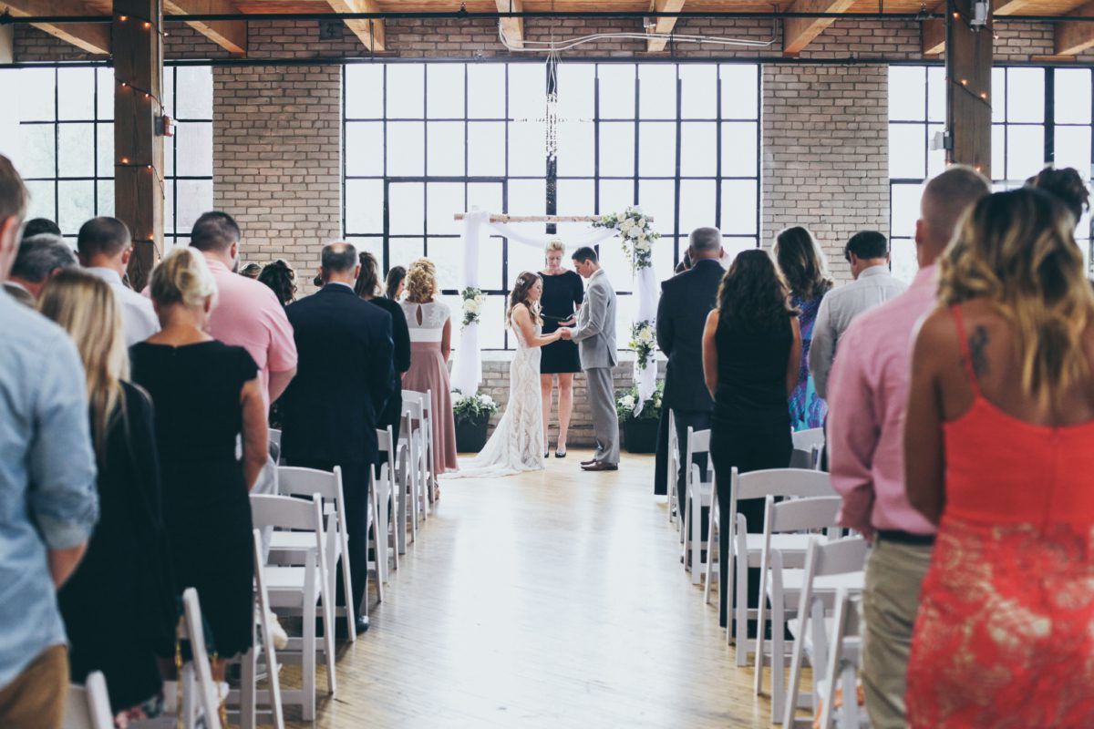 Wedding ceremony: Couple at altar; guests seated in rows. Industrial building with windows and exposed beams.