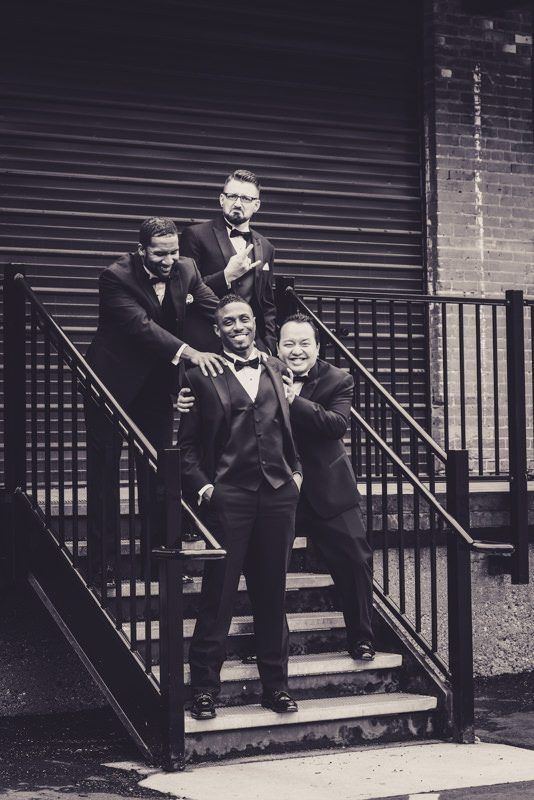 Four men in tuxedos pose on outdoor stairs near a brick building and a closed metal door.