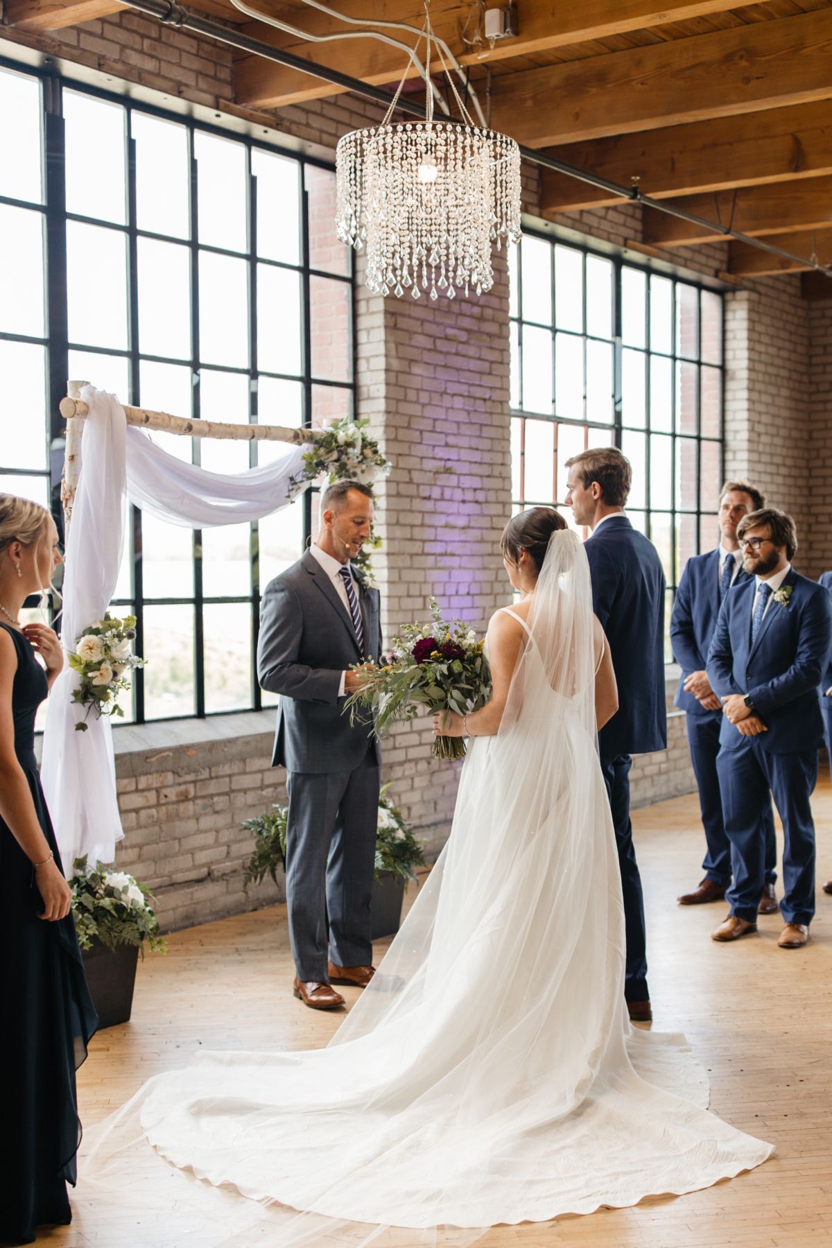 Wedding ceremony with bride, officiant, and groomsmen in a room with large windows.