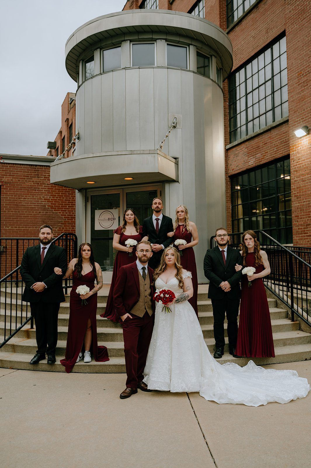 Wedding party on steps in front of a brick building. Bride and groom in center, bridesmaids in burgundy dresses, groomsmen in suits.