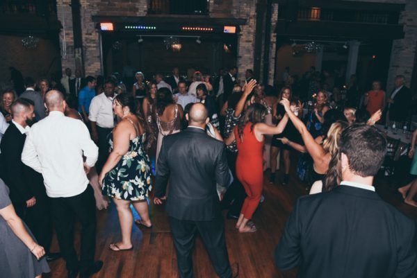 People dancing in a brightly lit room with a wooden floor, brick walls, and overhead lighting.