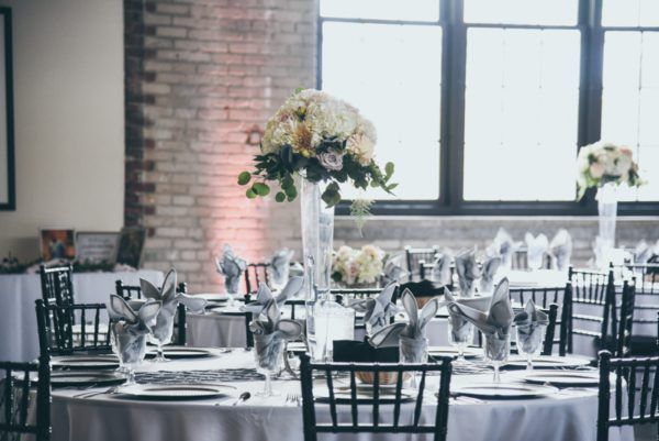 Round tables set for a wedding reception in a room with brick walls and large windows, floral centerpieces.