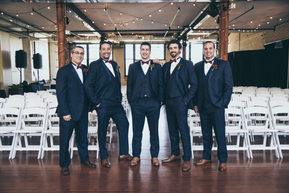 Five men in navy suits, bow ties, and brown shoes pose in a ceremony hall with white chairs.