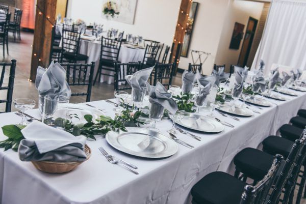 Long banquet table set for a formal event with white linens, silver accents, and greenery.