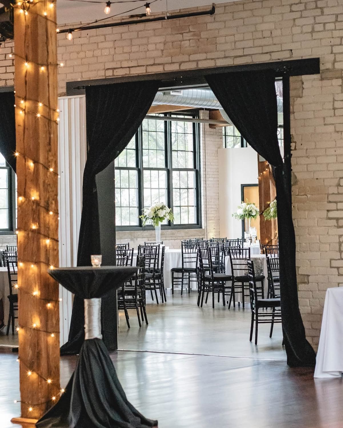 Event space with brick walls, open doorway, tables, chairs, and black draped curtains.