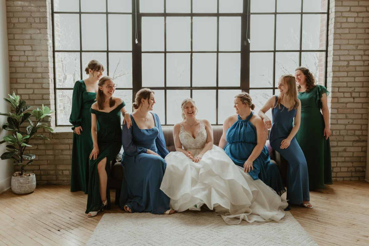 Bride with bridesmaids in blue and green gowns poses by a window, laughing.