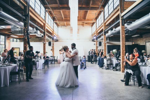 Couple dancing in large industrial space at a wedding reception; guests seated at tables.