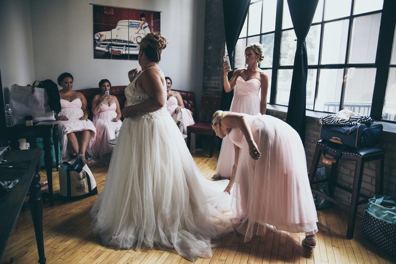 Bride in wedding dress, attendants in pink, preparing in a room with a large window.