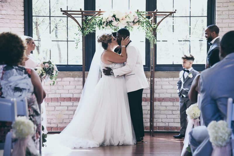 Bride and groom kiss at altar during ceremony. Wooden arch adorned with flowers; guests seated.