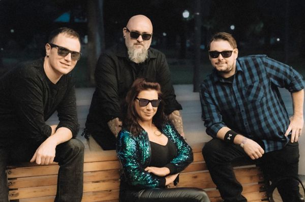 Band of four posing outdoors at night, wearing sunglasses; woman in sequin jacket.
