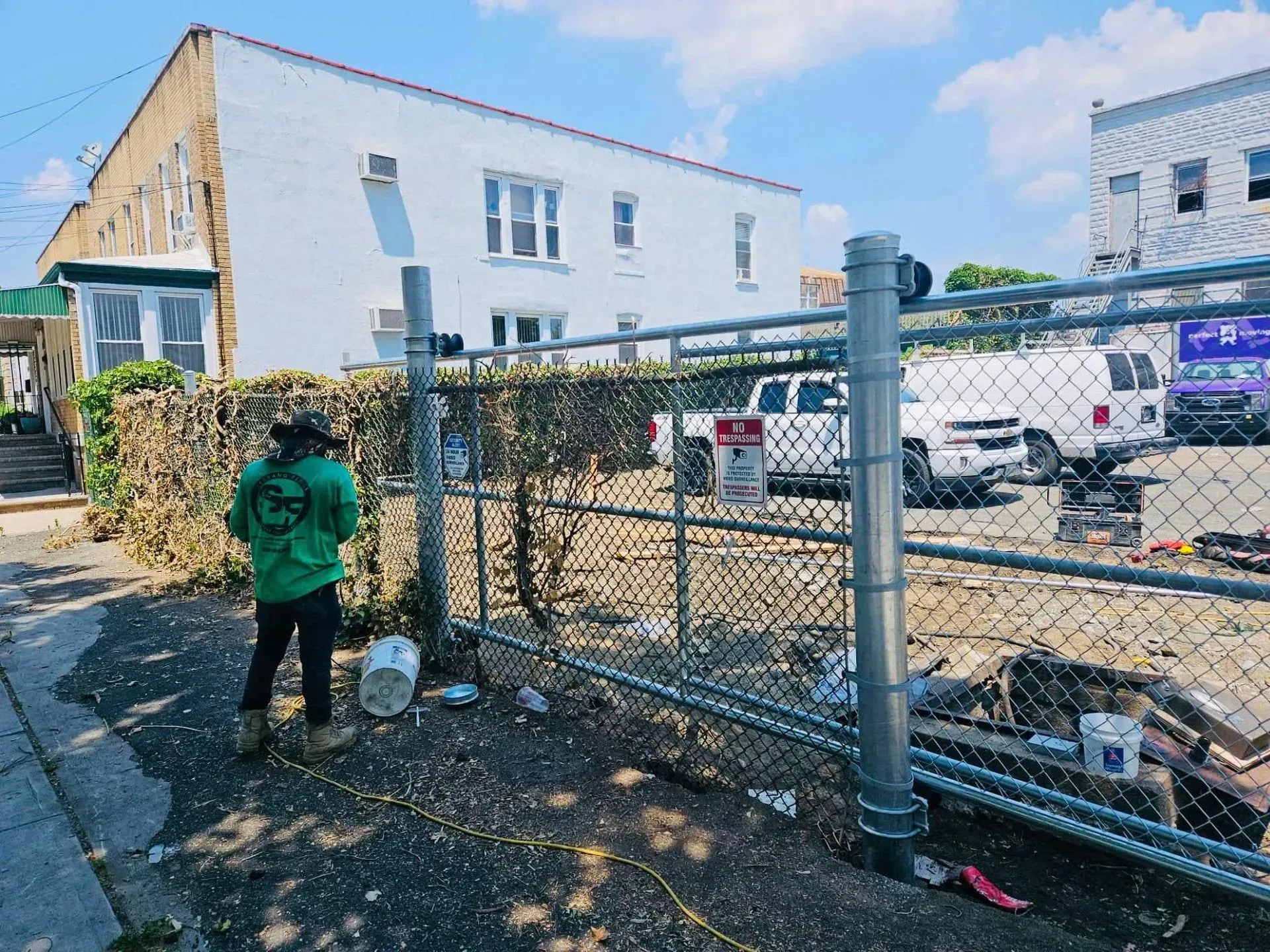 Person in green shirt by a chain-link fence, white building in the background, construction site.