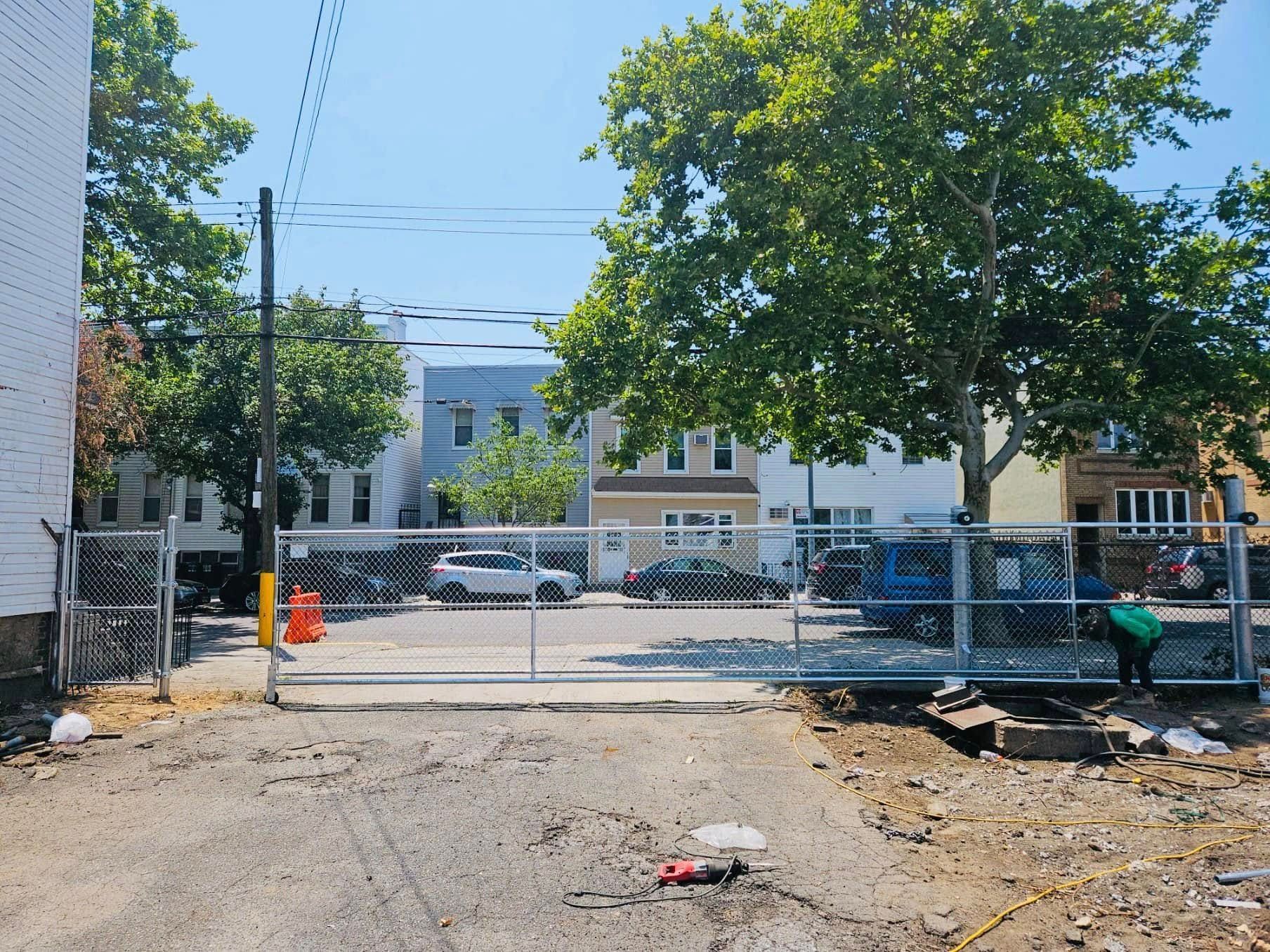 Gated lot with chain-link fence, parked cars, and a worker. Buildings and trees are in the background.