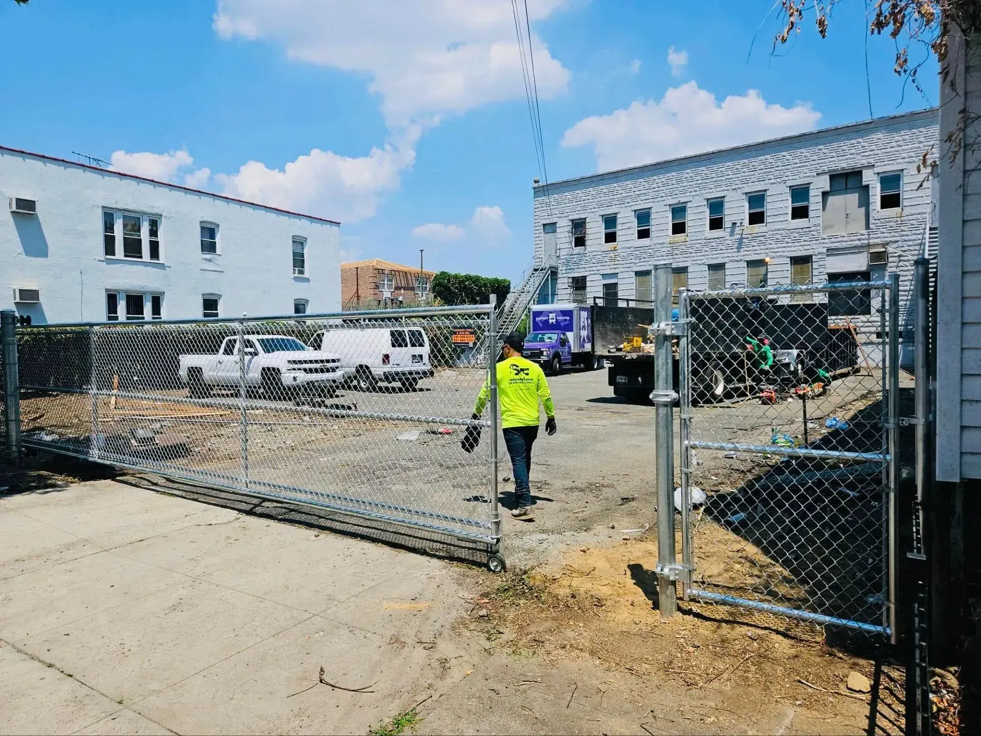 A person walks towards a chain link fence-enclosed construction site with vehicles. Sunny day.