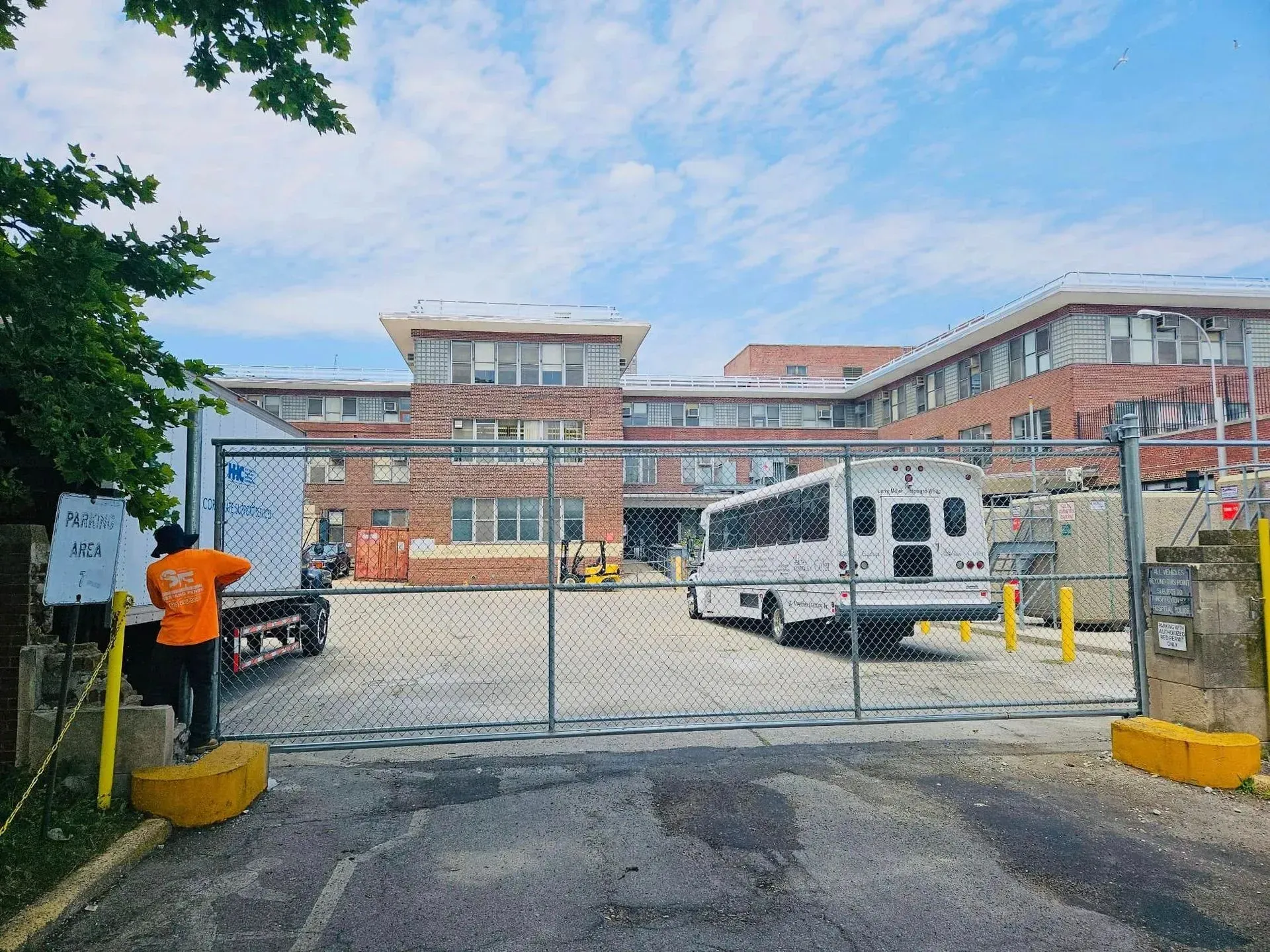 A gated brick building with a bus and construction equipment. A person in orange stands near the fence.