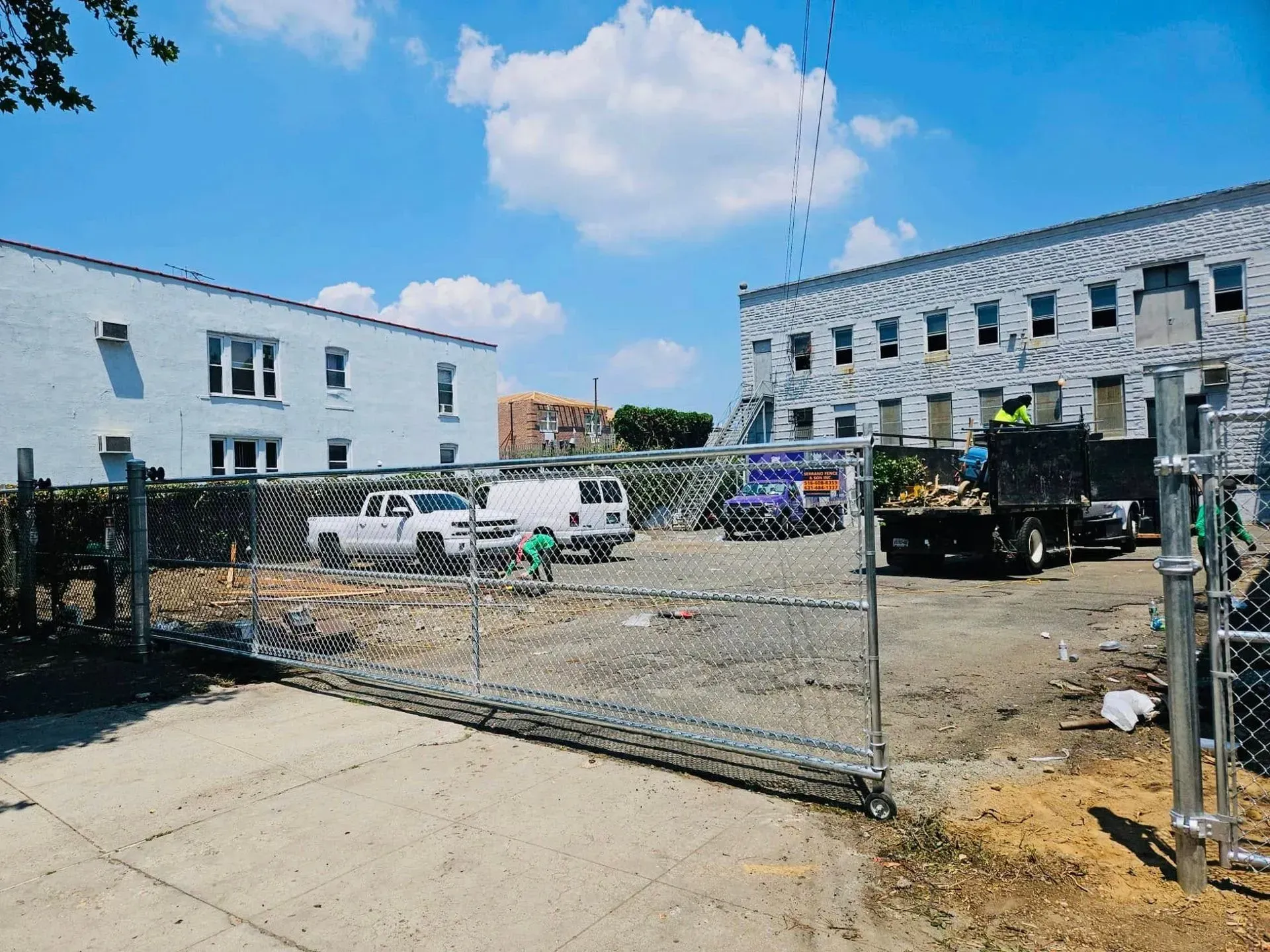 Construction site with chain-link fence, white vehicles, and buildings under a blue sky with clouds.