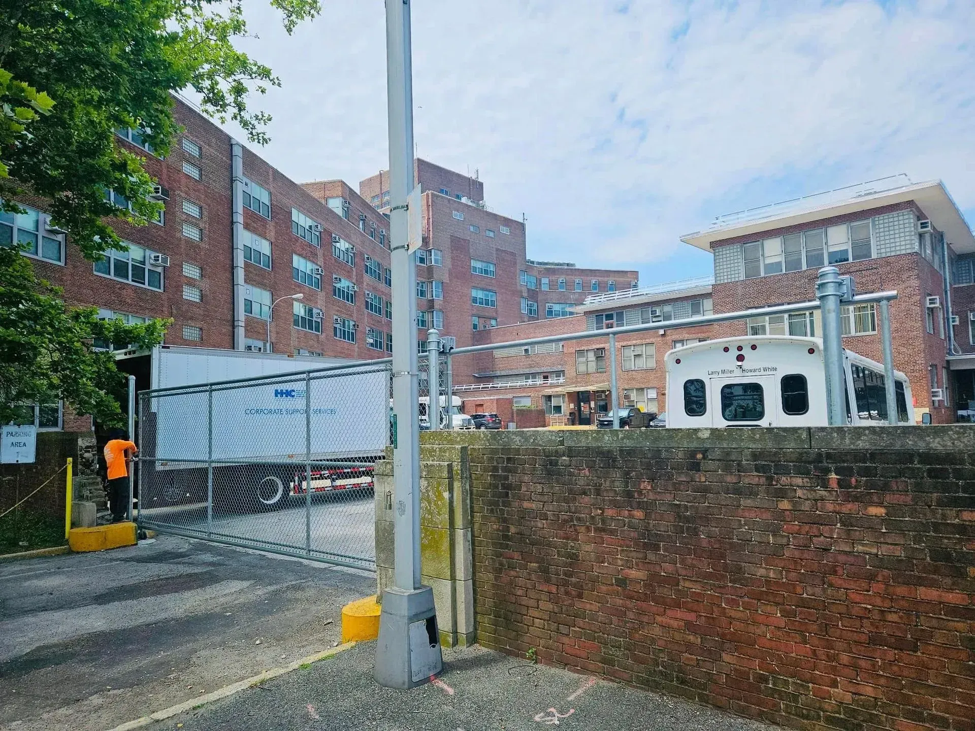 Brick building and gate, with a worker in orange vest. Blue sky with some clouds.