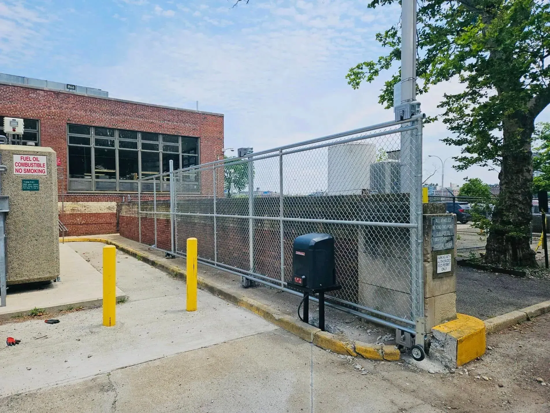 Chain link fence and gate enclosing area near a brick building. Yellow bollards and concrete pavement.