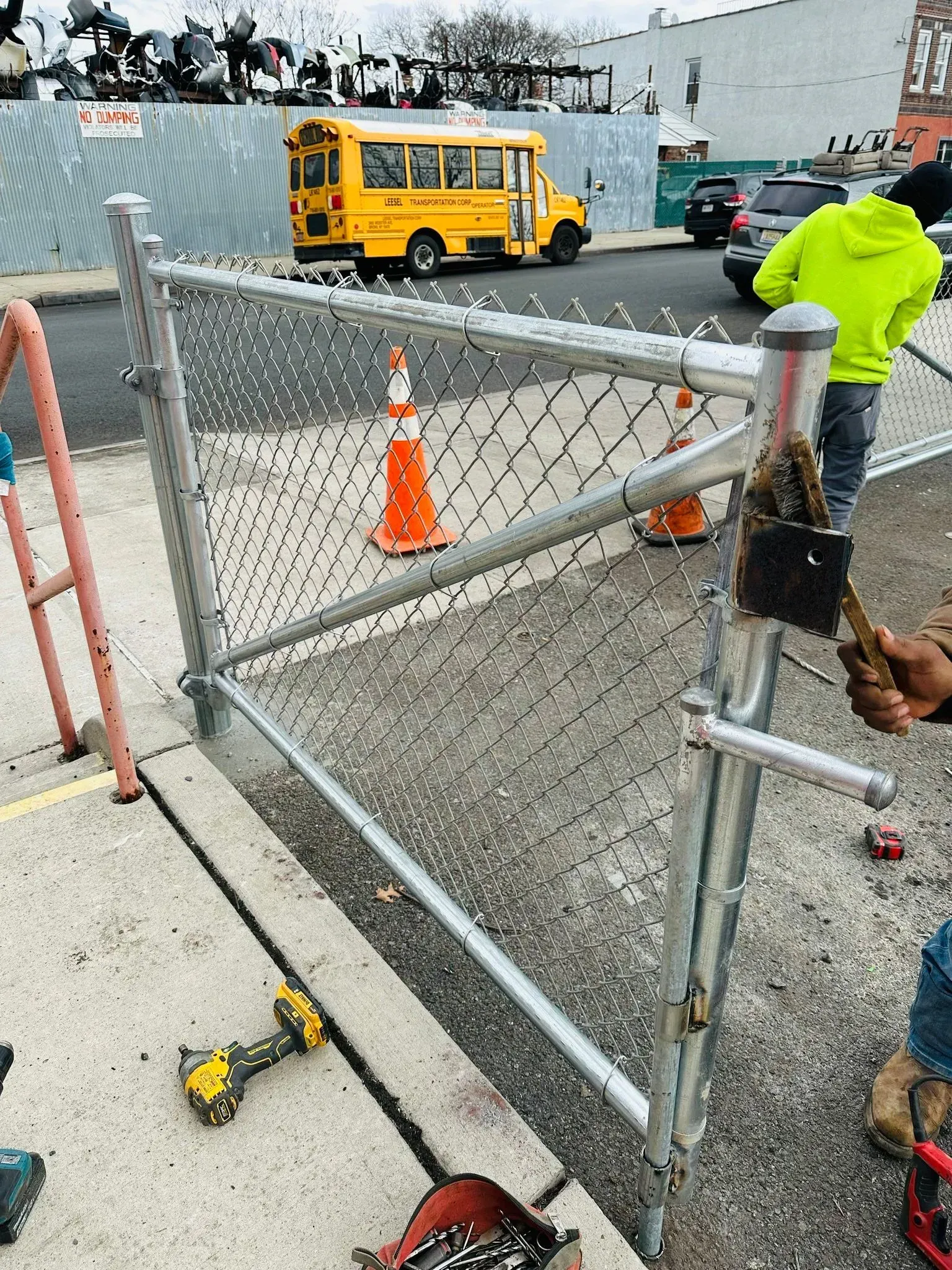 Chain-link fence gate being worked on with tools on the ground. A school bus and a worker wearing a neon green jacket are in the background.