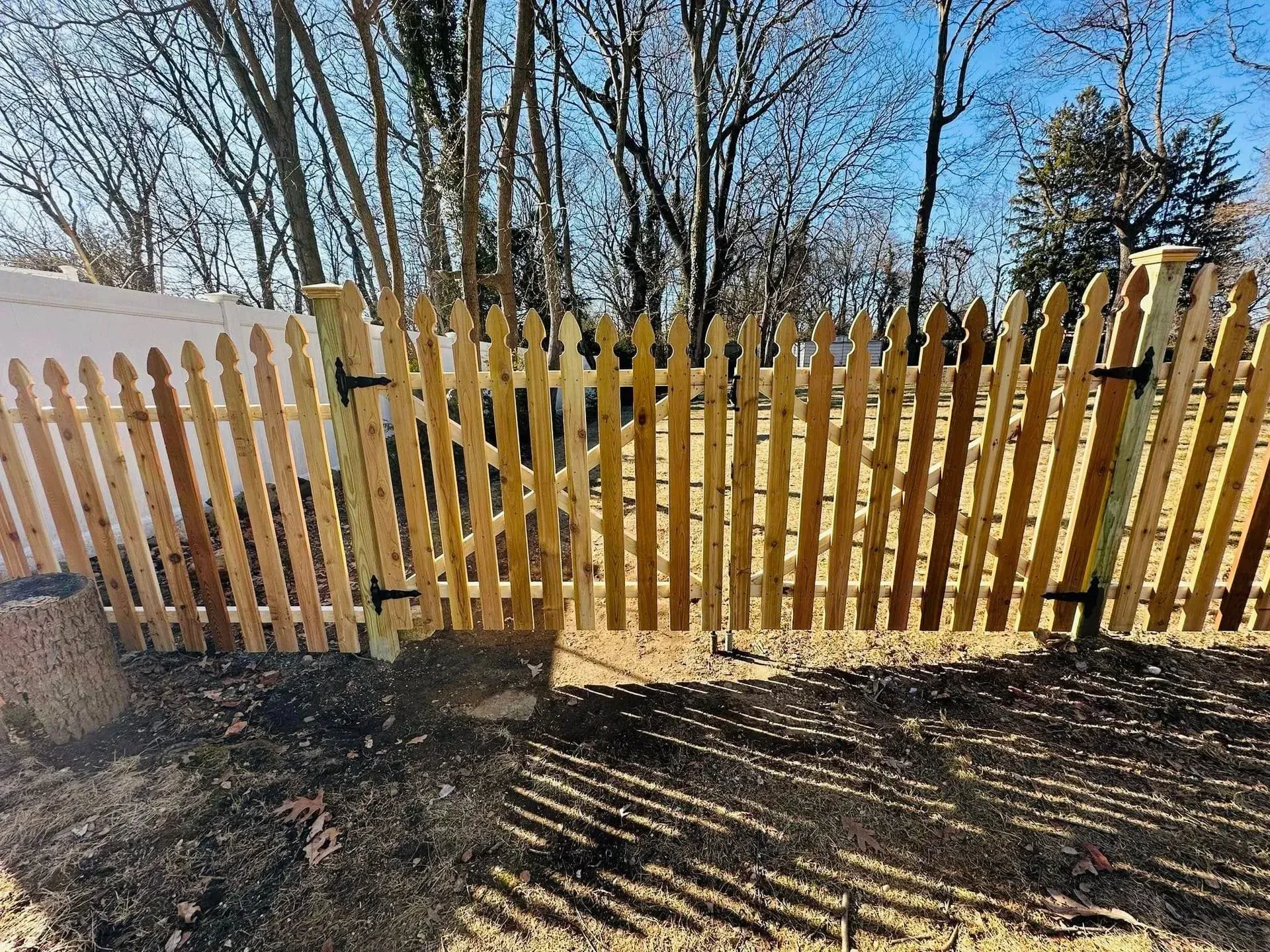 Wooden picket fence with gate, outdoors. Brown pickets, black hinges, dirt ground, trees, blue sky.