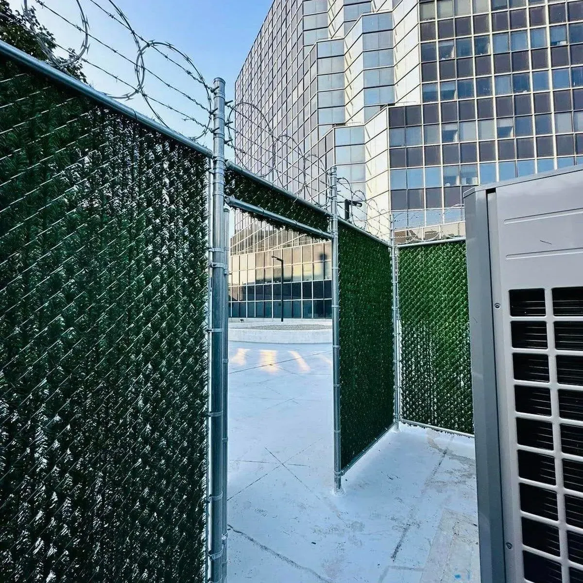 Chain-link fence with green privacy screening and barbed wire on a rooftop with a building in the background.