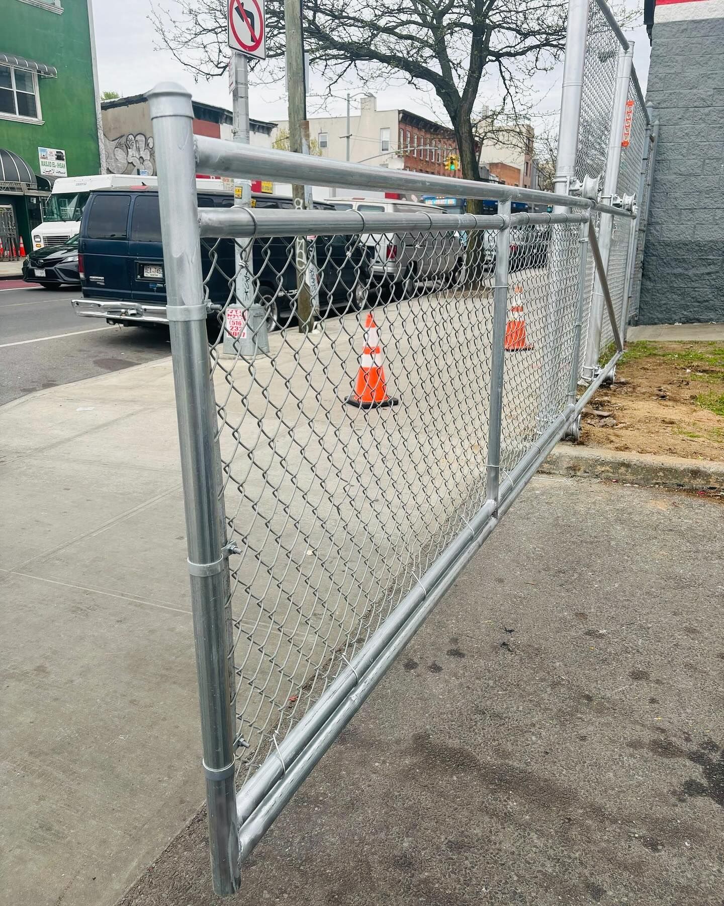 Chain-link fence on a sidewalk, adjacent to a street with cars and orange cones.
