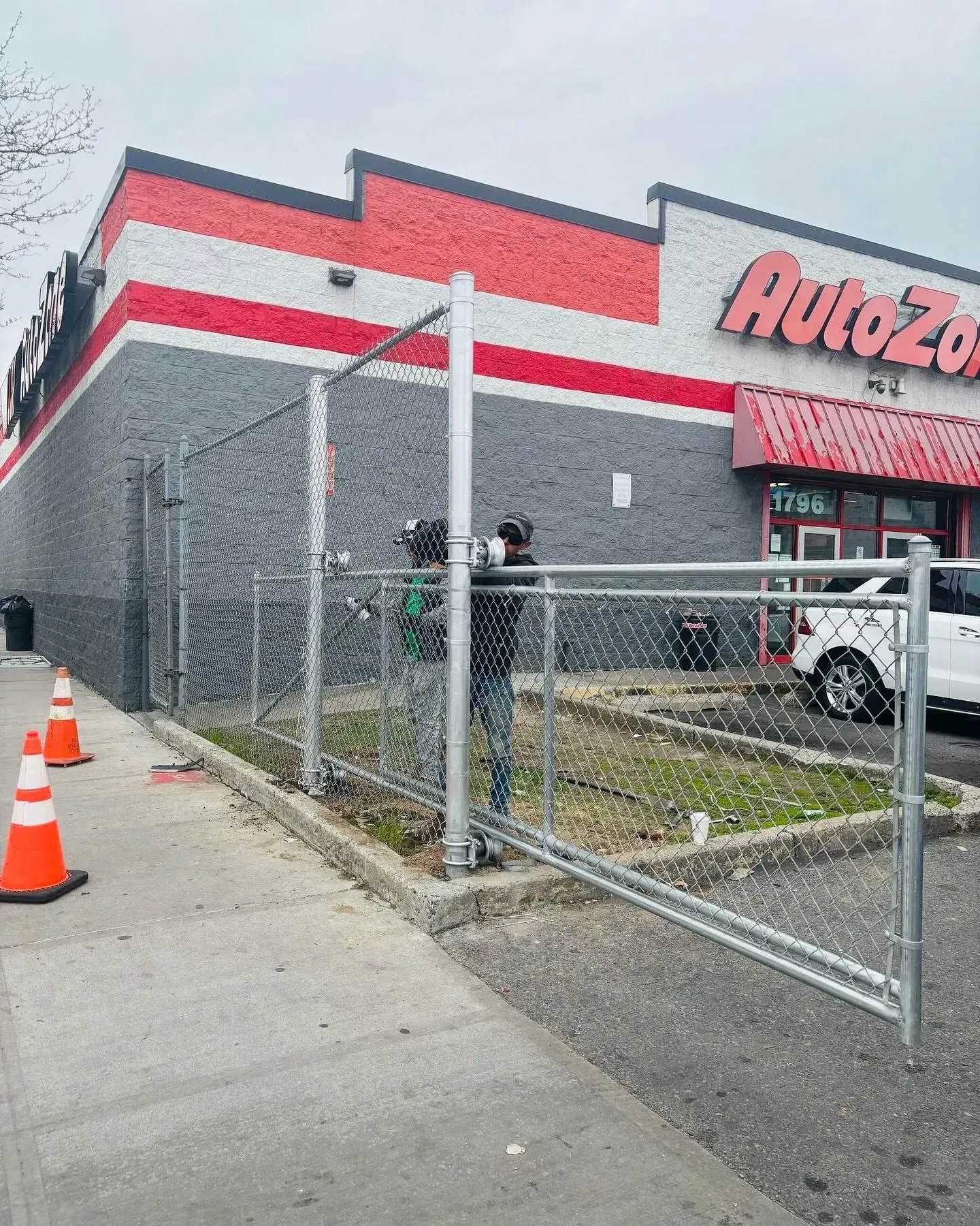 AutoZone store with a chain-link fence being constructed in front. Gray and red building, orange traffic cones.