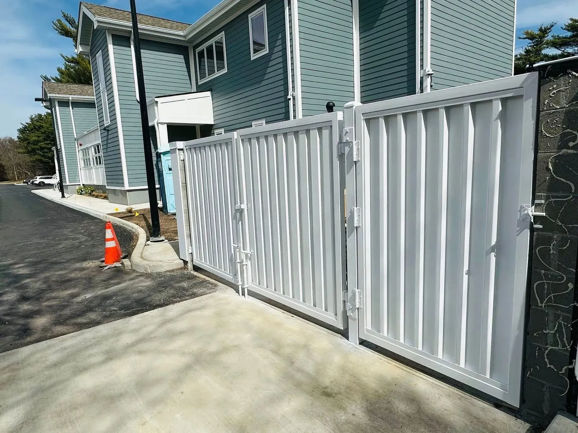White metal gate in front of a blue building, on a paved area with a traffic cone.