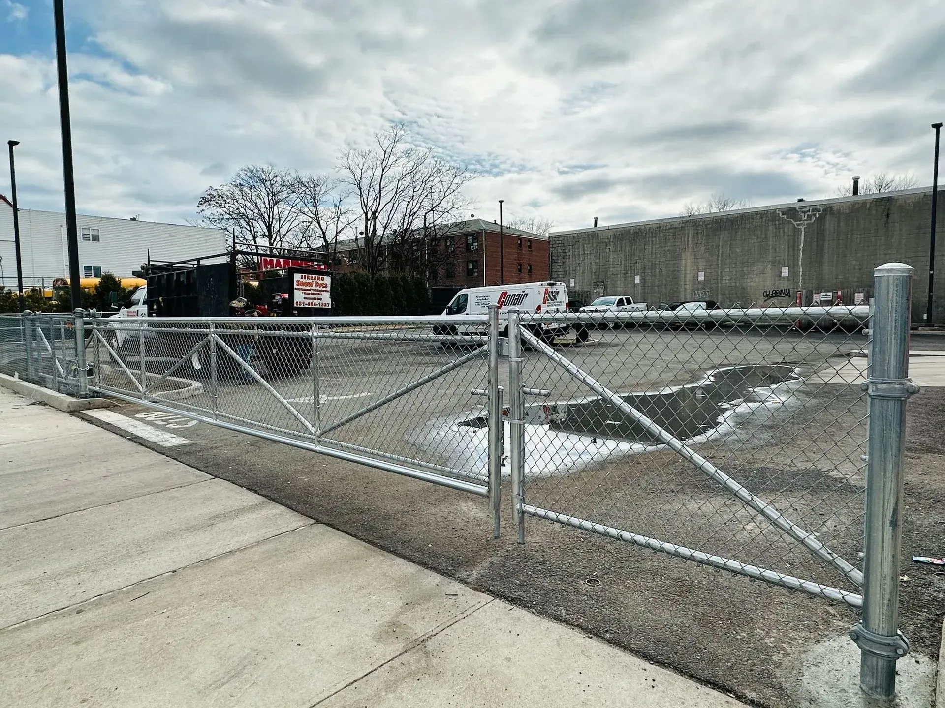 Chain-link fence bordering a paved area and a muddy gravel lot under a cloudy sky.
