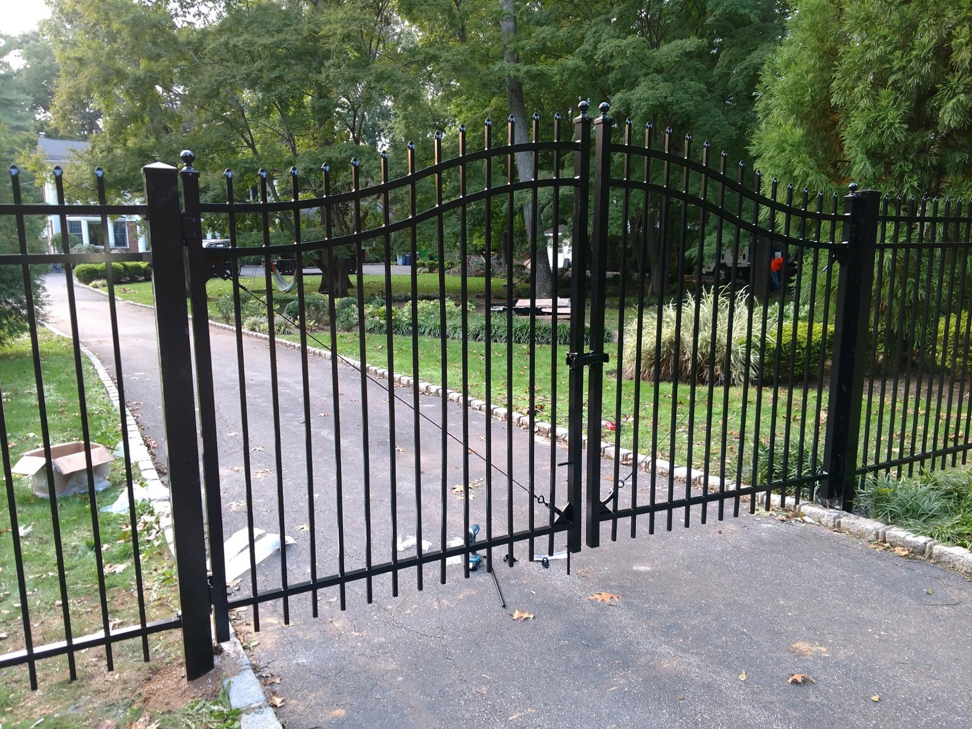 Black metal driveway gate, open, leading to a yard with trees.