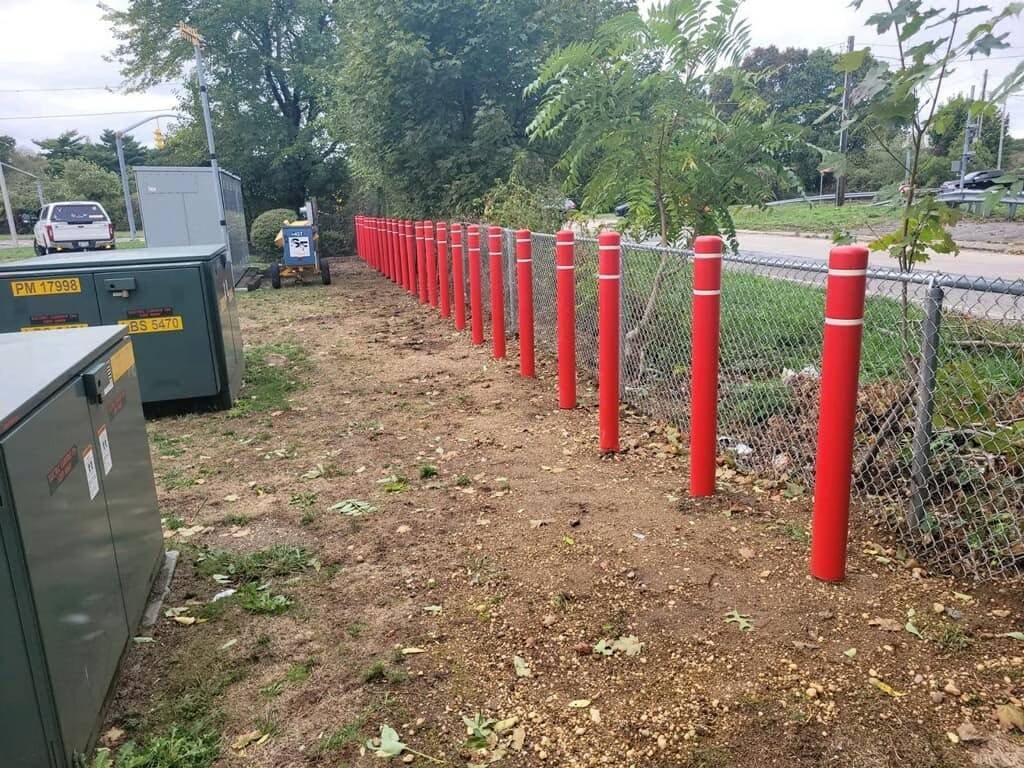 Red bollards line a dirt area next to a chain-link fence and green utility boxes.