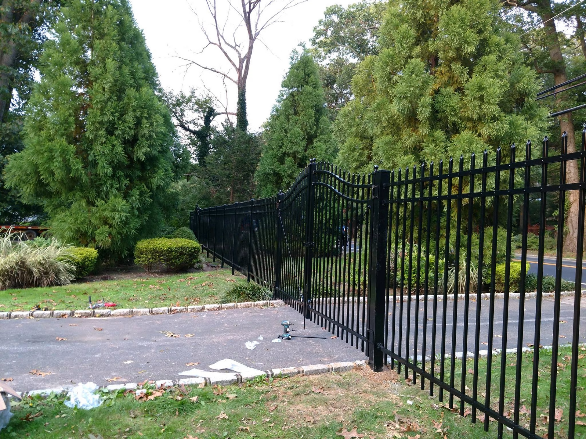 Black metal fence along a driveway with trees and grass in the background.