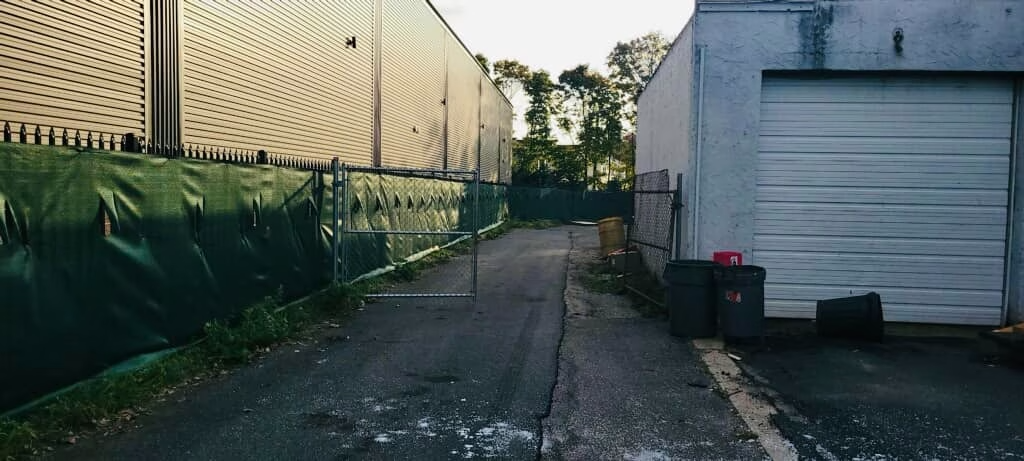 A narrow, cracked, asphalt alley between a building with a closed garage door and a patterned wall.