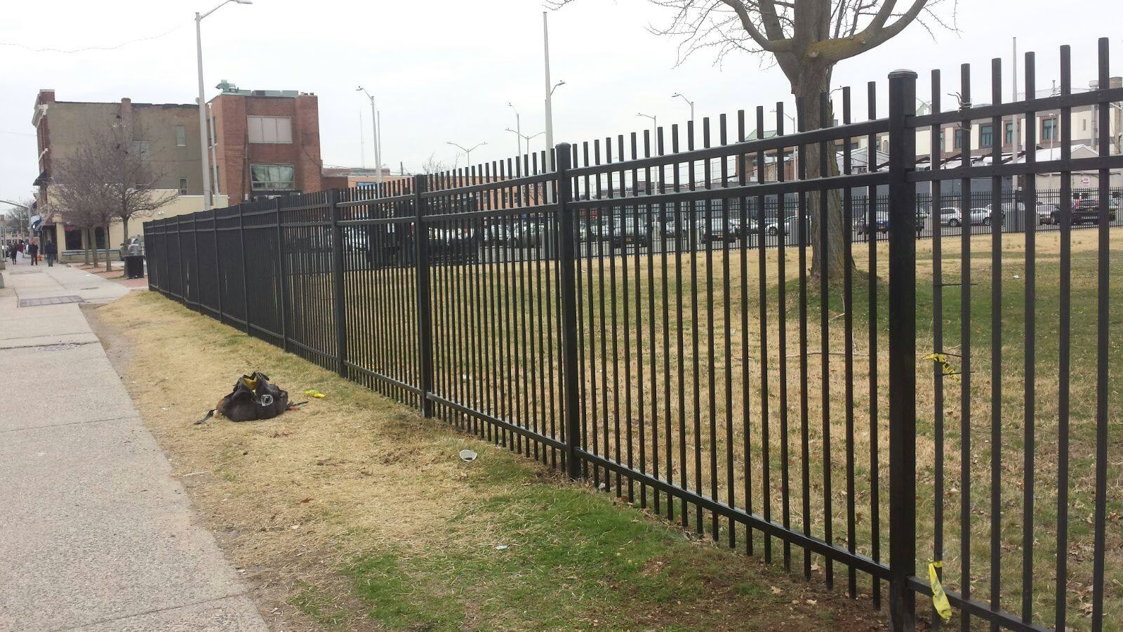 Black metal fence bordering a grassy area and sidewalk. Buildings and a tree are in the background.