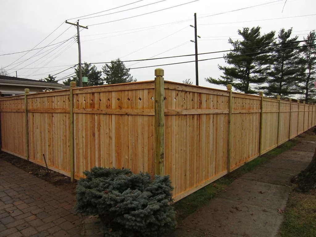 Wooden privacy fence in a residential area, corner view. Cloudy sky, evergreen bush, and sidewalk.