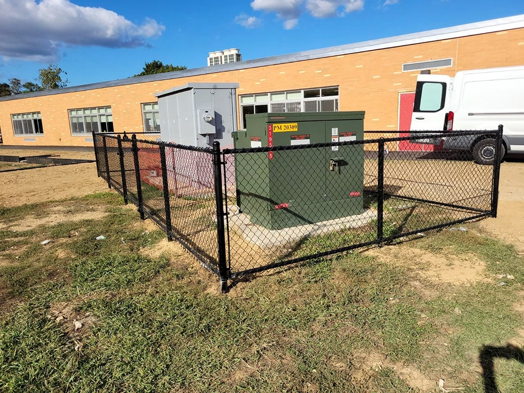 Black chain-link fence encloses electrical equipment near a red-brick building with a white van in the background.
