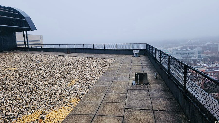 Rooftop view on a cloudy day. Gravel and concrete tiles on the roof; metal railing surrounds. City buildings in the distance.