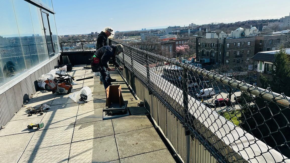 People on a rooftop working near a chain-link fence, overlooking a city.