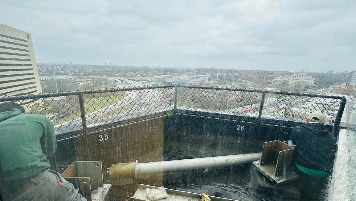 Workers on a high rooftop installing equipment; city view behind them with rain streaks.