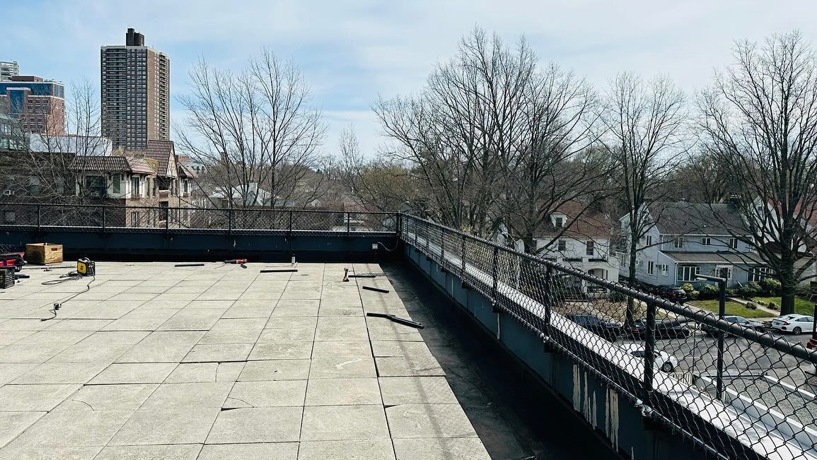 Rooftop view of a city with a low fence, buildings, and leafless trees under a blue sky.