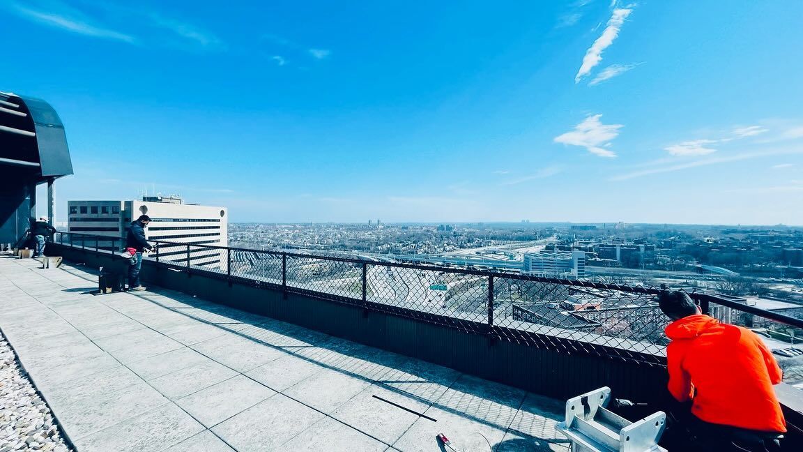 Cityscape view from a rooftop. Person in orange shirt leaning against railing, overlooking buildings on a clear day.