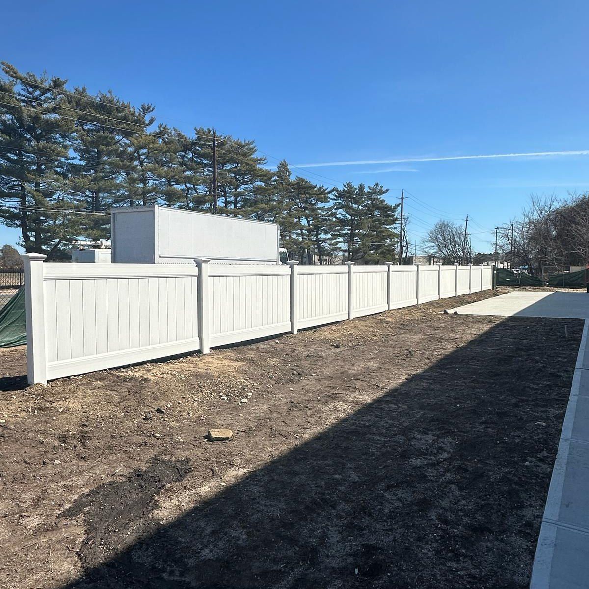 White vinyl fence along a dirt area under a blue sky, with trees and building visible.
