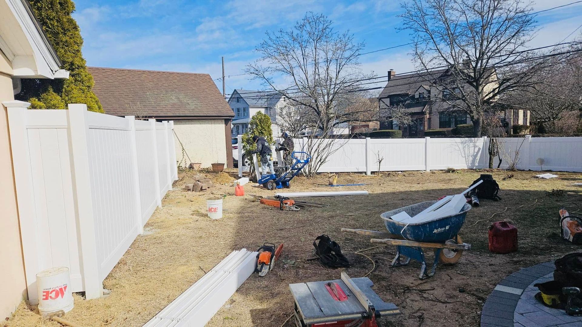 Backyard with white fence, tools, and construction materials on the grass.