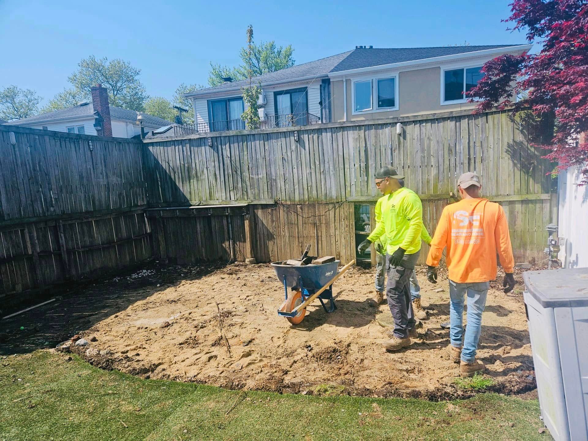 Two workers in safety vests clearing a backyard, wheelbarrow in the middle. Brown soil, wooden fence, sunny day.