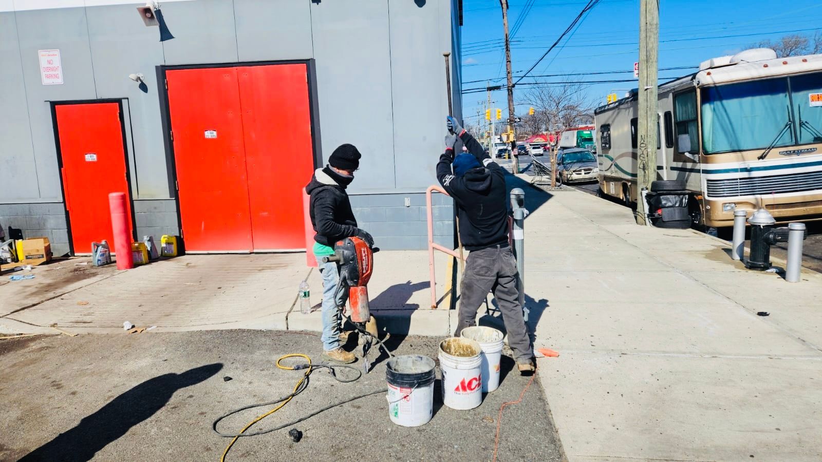 Two workers using tools to work on concrete near a red door and a parked RV on a sunny day.