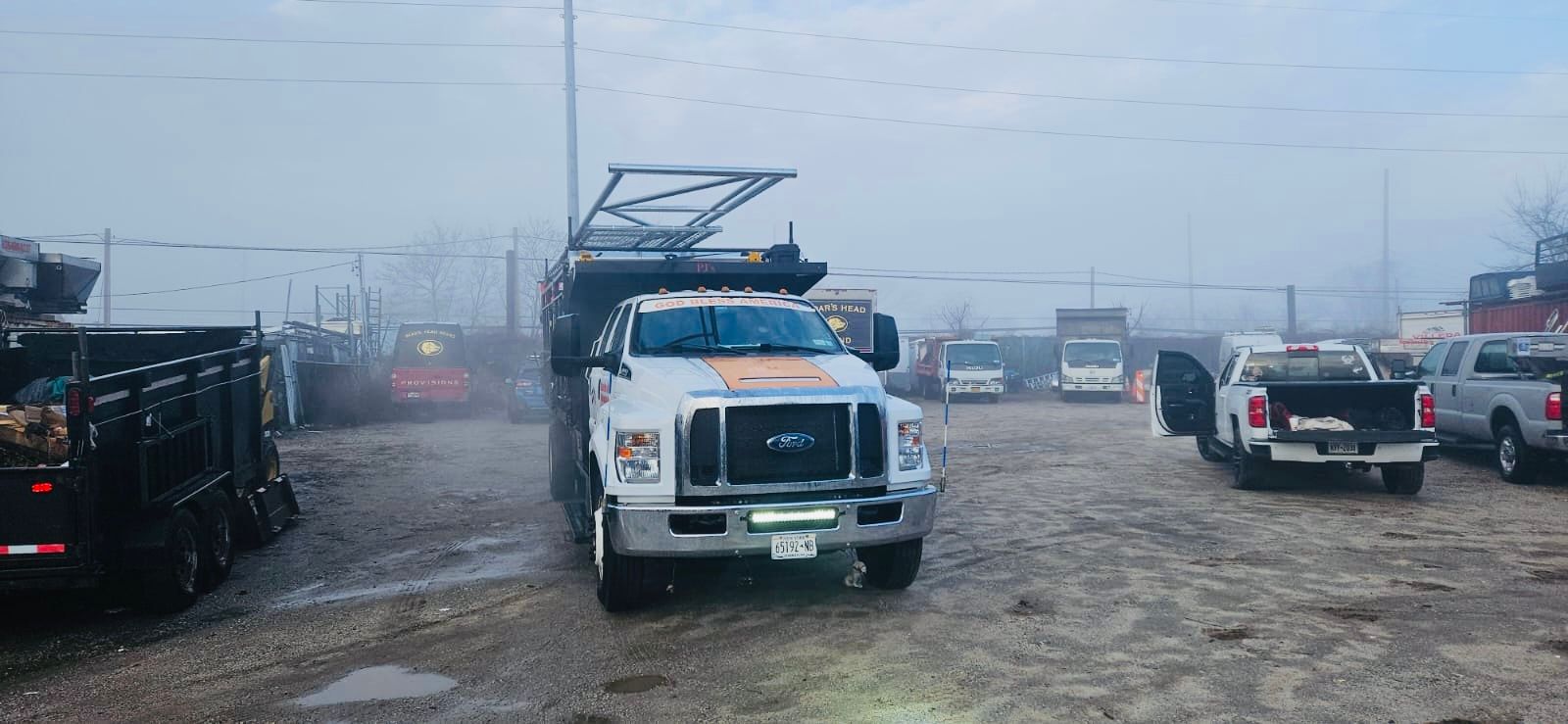 A white dump truck in a muddy lot with other trucks, under a hazy sky.
