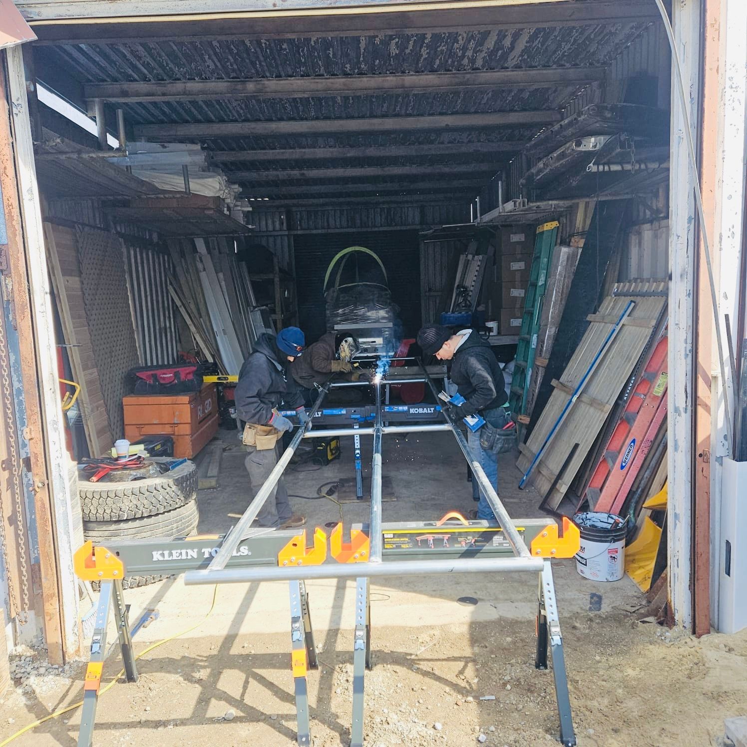 Two people working on a vehicle inside a cluttered garage. They are using a metal frame.