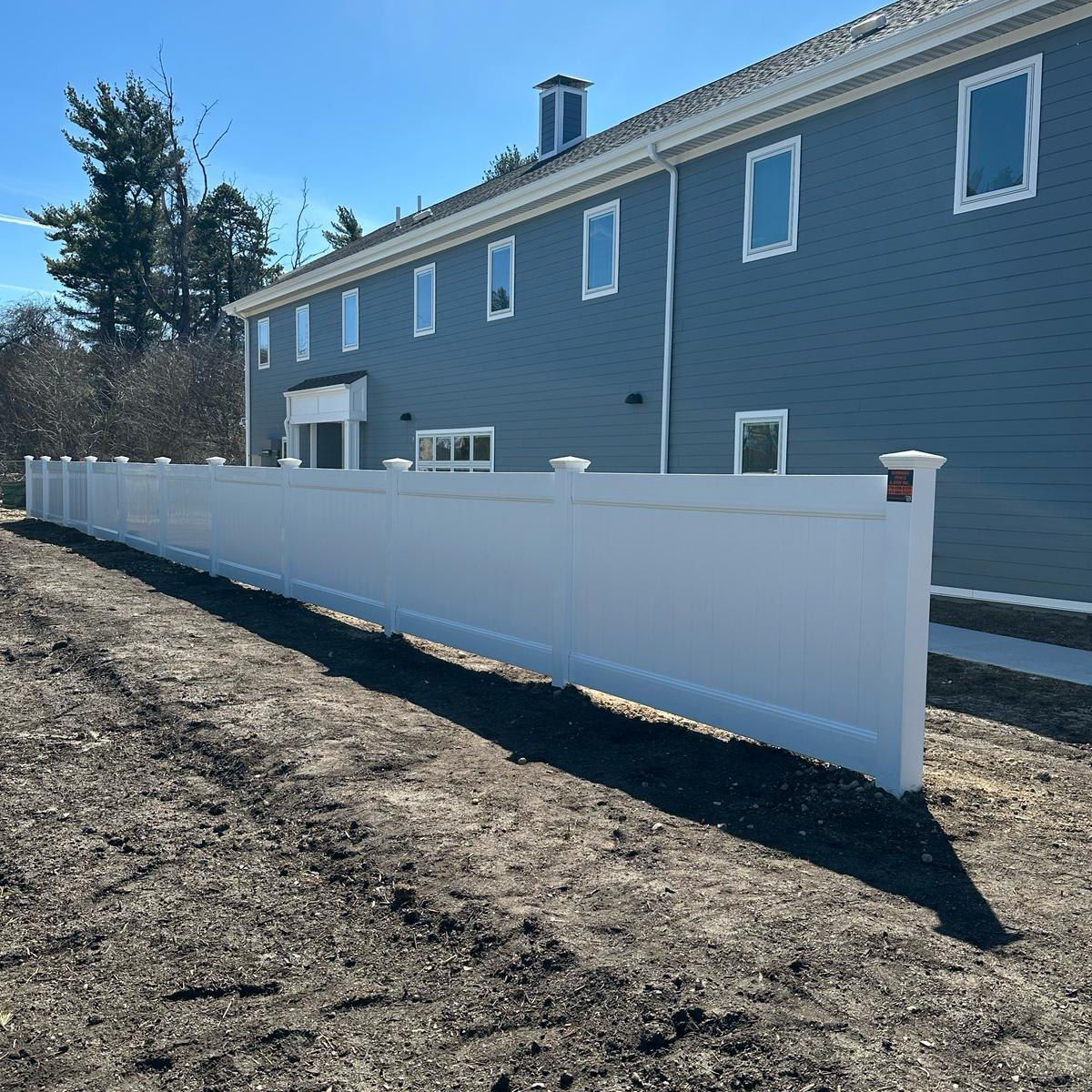 White vinyl fence along a blue-sided building with windows, set in a dirt yard. Sunny day.