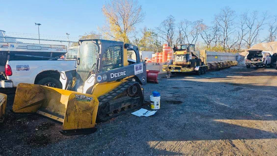 John Deere track loader with blade, parked with other construction vehicles and supplies outdoors.