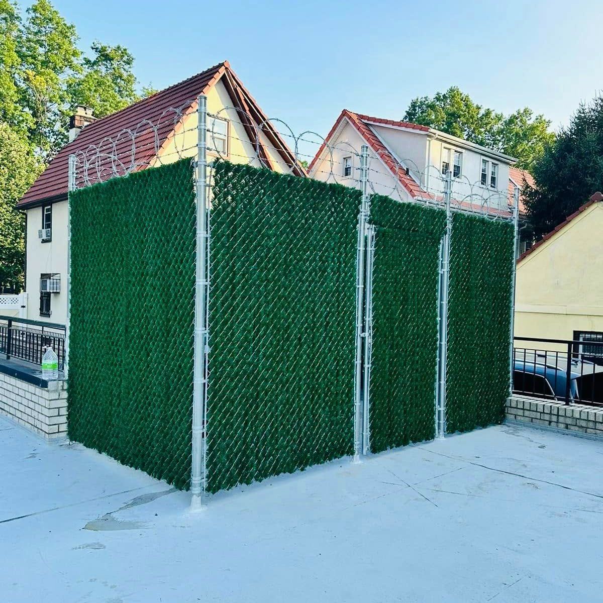 Green privacy fence with faux foliage and barbed wire, adjacent to a rooftop near houses.